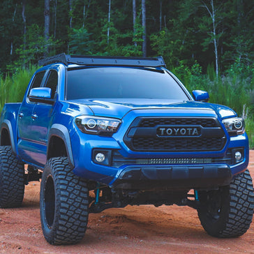 Lifted blue 3rd Gen Toyota Tacoma with a roof rack parked on a dirt trail in the woods